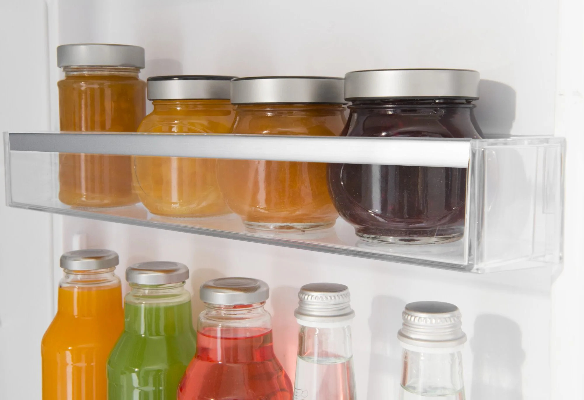 Półka w lodówce ze szklanymi słoikami i butelkami. Słoiki zawierają dżem, butelki sok. Kolory: pomarańczowy, zielony i czerwony.