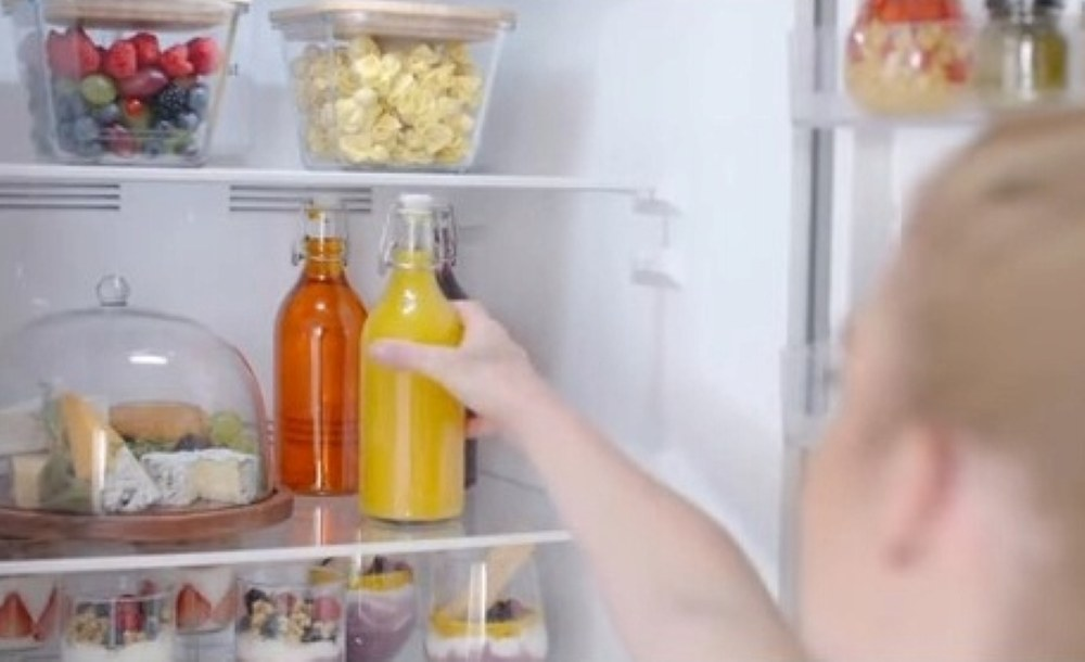 Ręka sięga do lodówki. Widoczne są słoiki z owocami i sokiem. W środku jest też ser i desery.