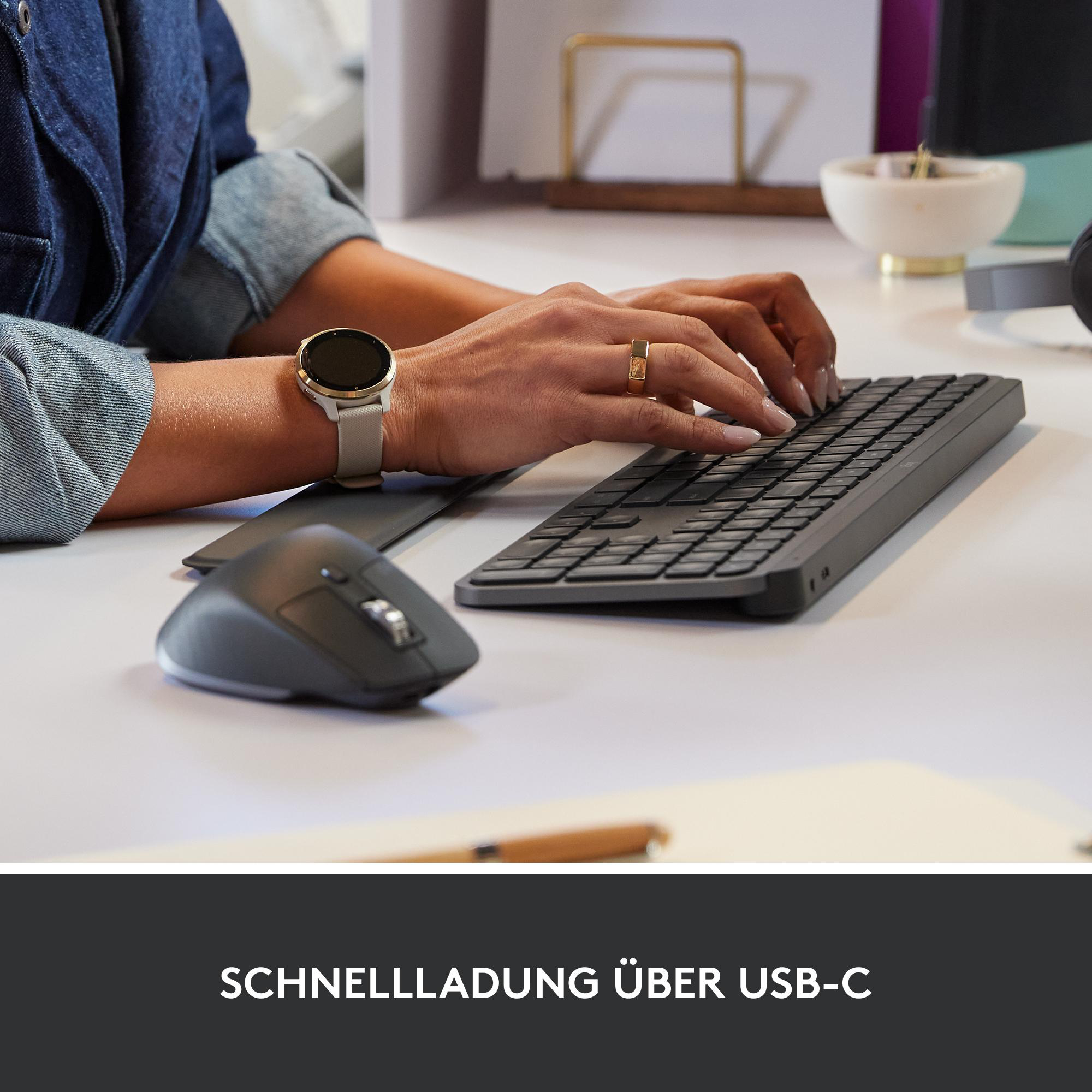 Hände tippen auf einer schwarzen Tastatur. Eine Armbanduhr, ein Ring und eine Maus sind sichtbar. Der Hintergrund ist ein weißer Schreibtisch.