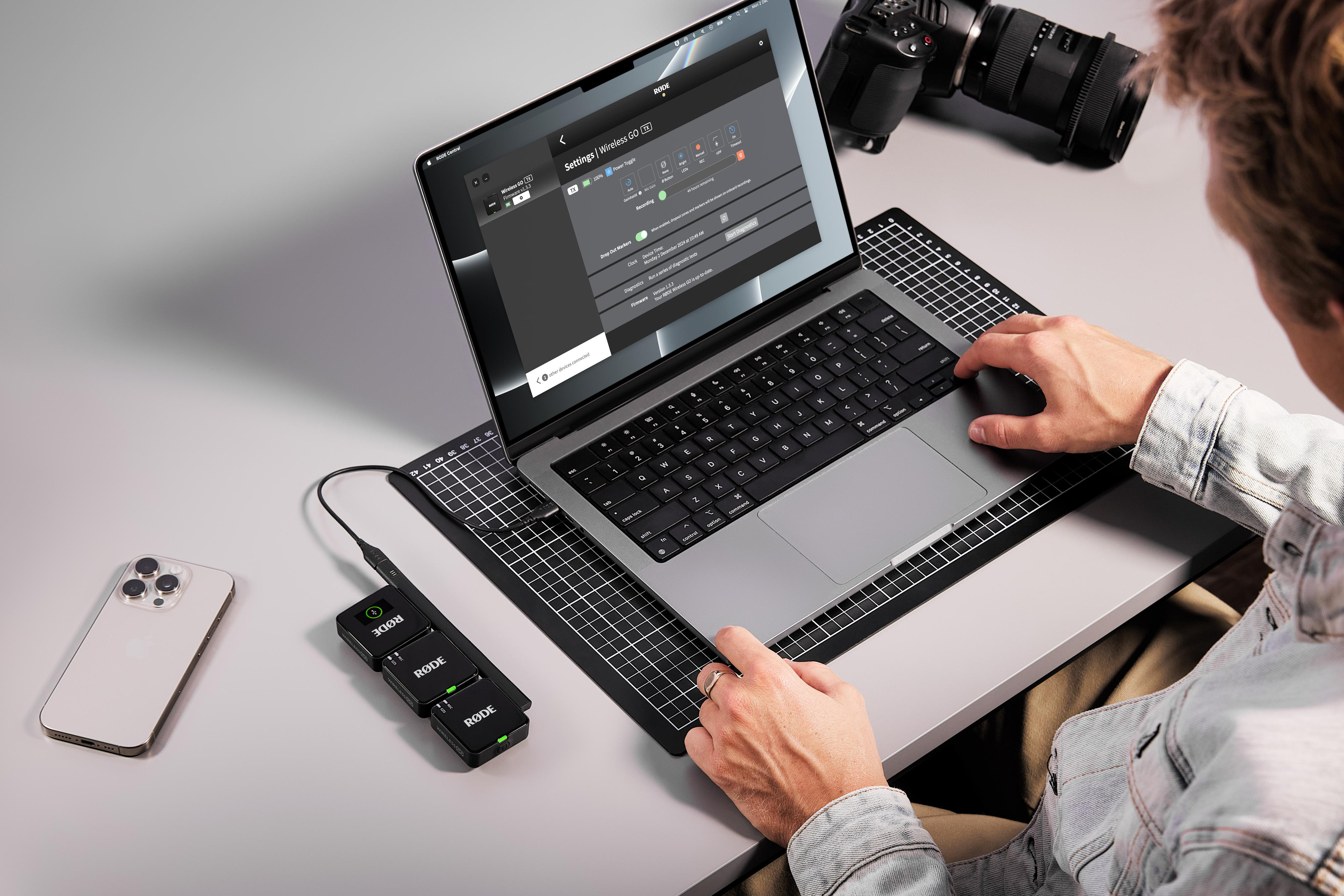 Eine Person nutzt einen Laptop mit Audioausrüstung in der Nähe. Der Bildschirm zeigt Einstellungen. Ein Telefon und eine Kamera sind ebenfalls sichtbar.