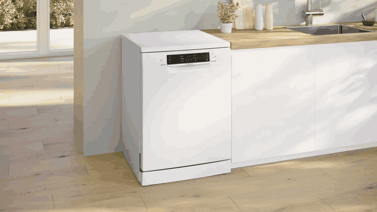 Un lavavajillas blanco se encuentra en una cocina. La cocina tiene encimeras de madera y armarios blancos. El suelo es de madera.