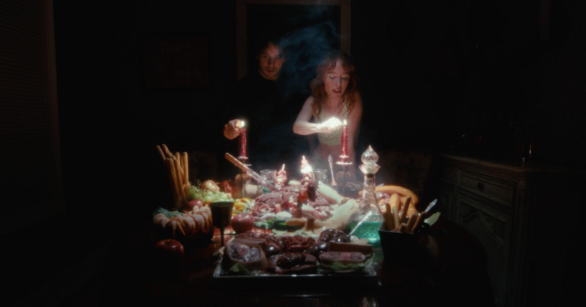 Zwei Personen zünden Kerzen über einem Tisch voller Essen an. Rauch steigt auf, die schwache Beleuchtung erzeugt eine geheimnisvolle Atmosphäre. Früchte und Fleisch sind sichtbar.