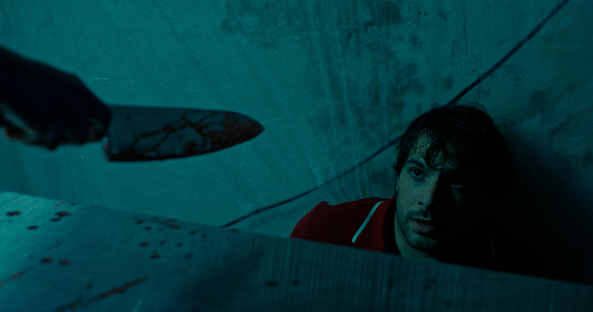 Ein blutiges Messer wird über einem Mann gehalten, der aufschaut und gefangen ist. Die Szene hat einen teal-Farbton, was auf einen Horrorfilm hindeutet.