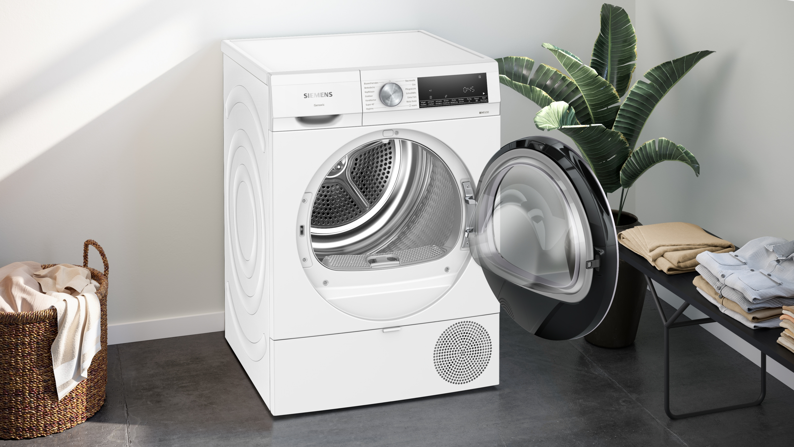 Ein weißer Siemens-Trockner mit offener Tür. Ein Korb mit Stoff und eine Pflanze stehen an den Seiten. Kleidung liegt auf einem schwarzen Tisch.