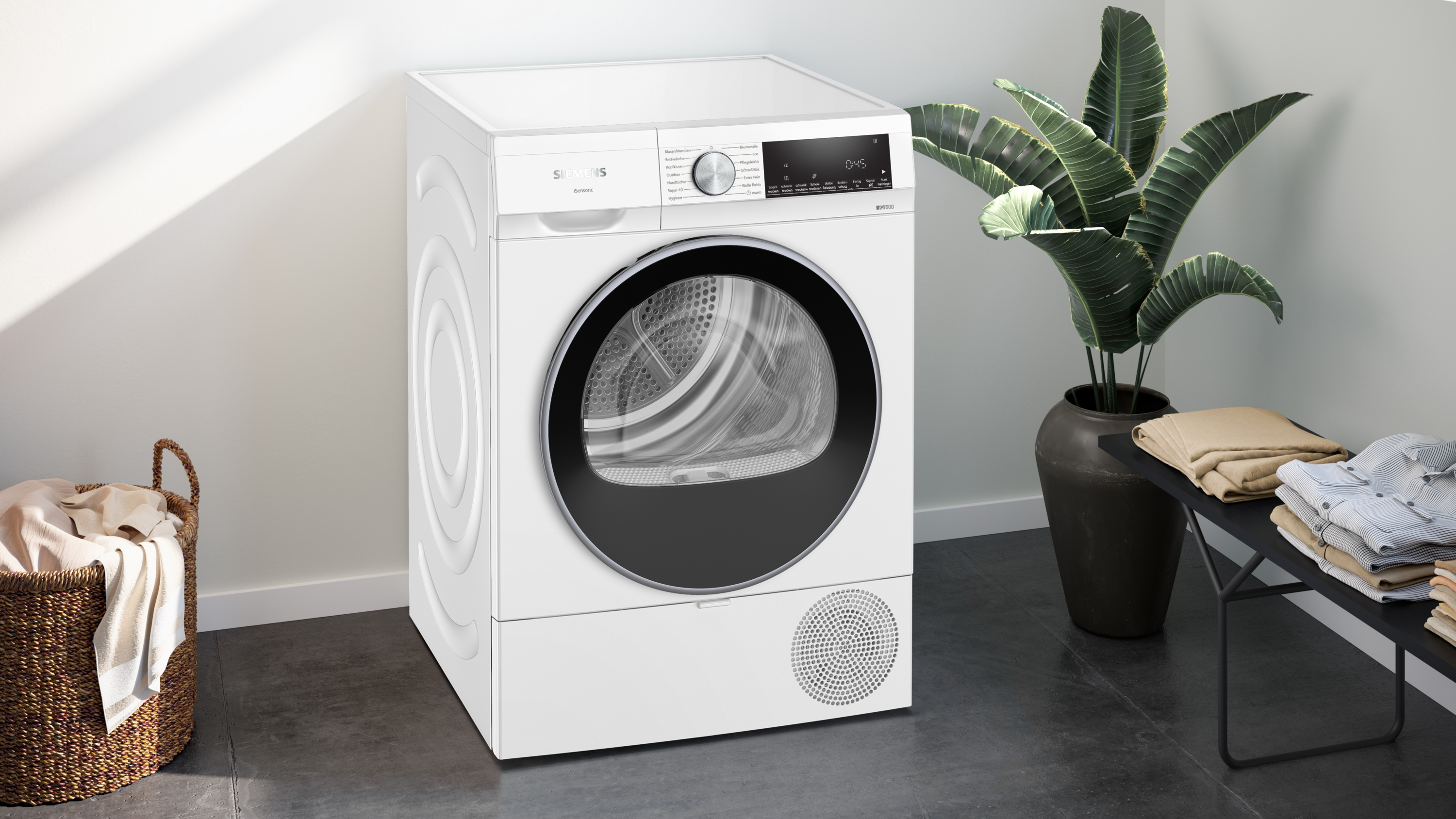 Ein weißer Siemens-Trockner steht neben einer Topfpflanze und einem Wäschekorb. Kleidung liegt auf einem Tisch.