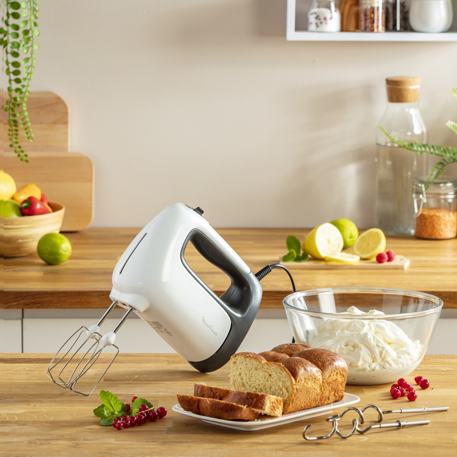 Una scena di cucina con un frullatore a mano bianco, pane, panna montata e frutta su una superficie di legno. Sono visibili bacche e limoni.