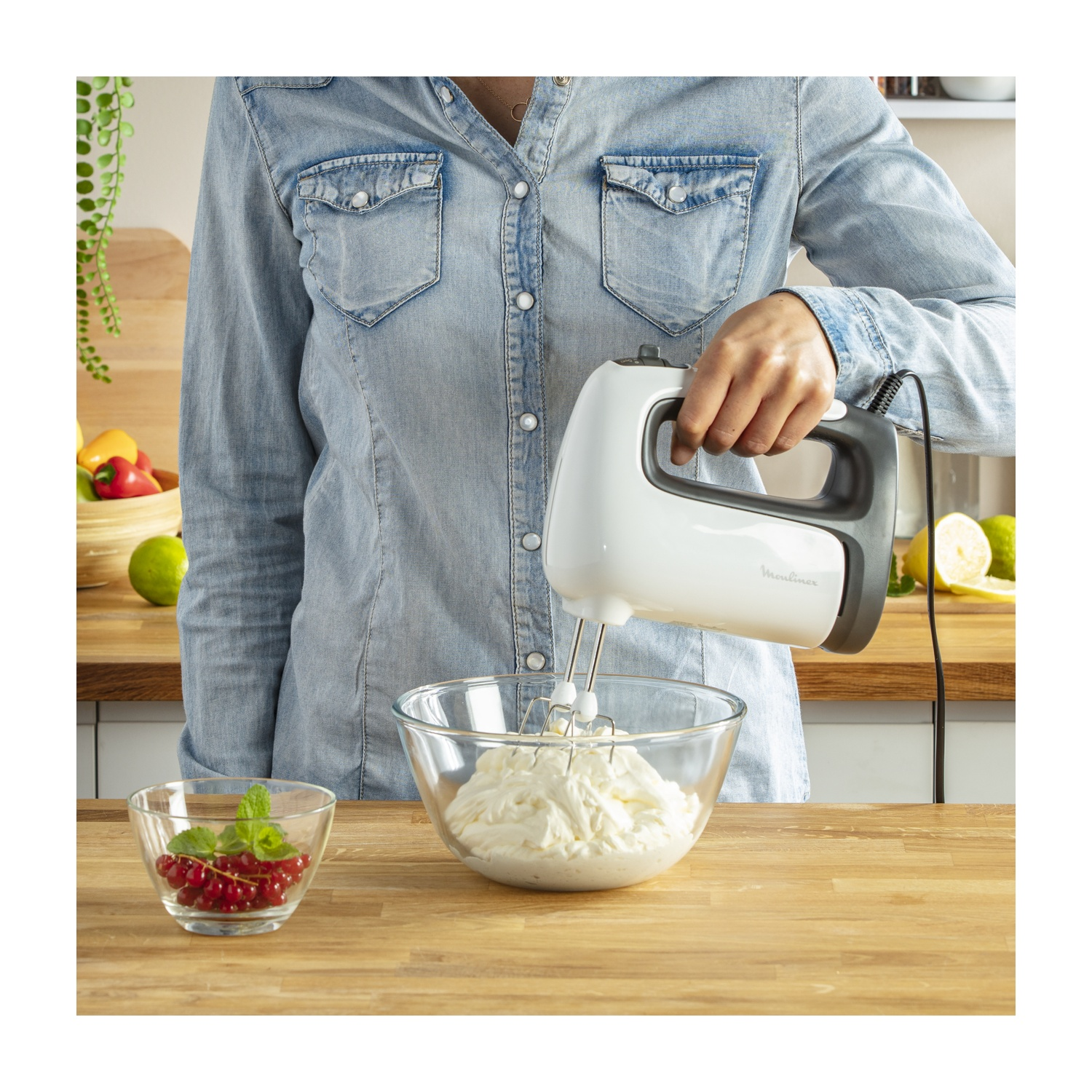 Una donna con una camicia di jeans usa un mixer bianco per montare la panna in una ciotola di vetro. Ribes rossi in una ciotola piccola sono sul bancone.