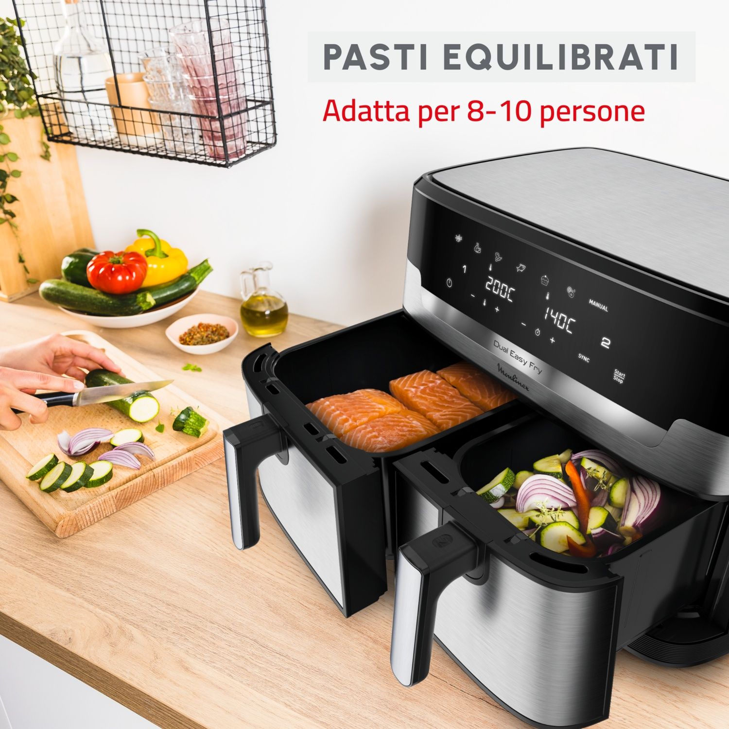 Una friggitrice ad aria con due scomparti. Uno ha salmone, l'altro verdure. Una persona taglia verdure su un tagliere.
