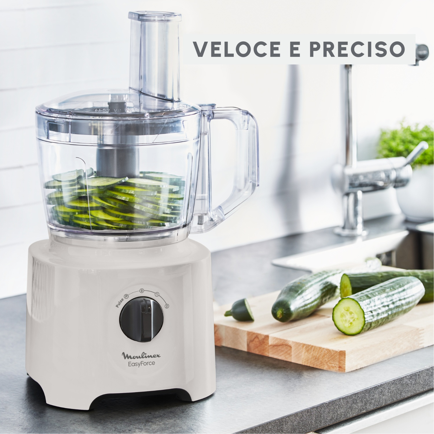 Un robot da cucina bianco con cetrioli a fette all'interno. Cetrioli interi e un tagliere sul bancone.
