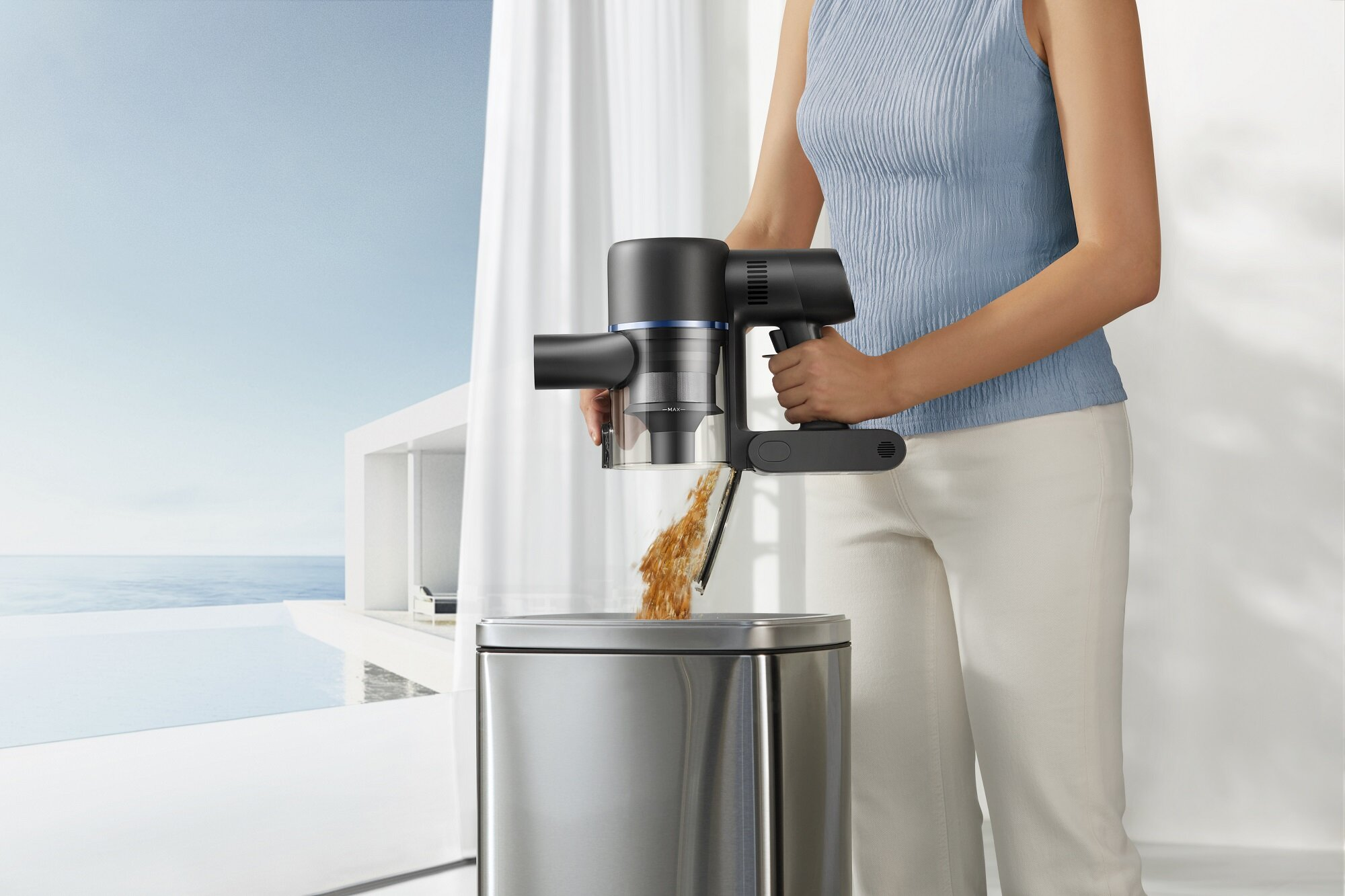 Kobieta opróżnia czarno-szary odkurzacz ręczny do srebrnego kosza na śmieci. W tle widać nowoczesny dom i morze.