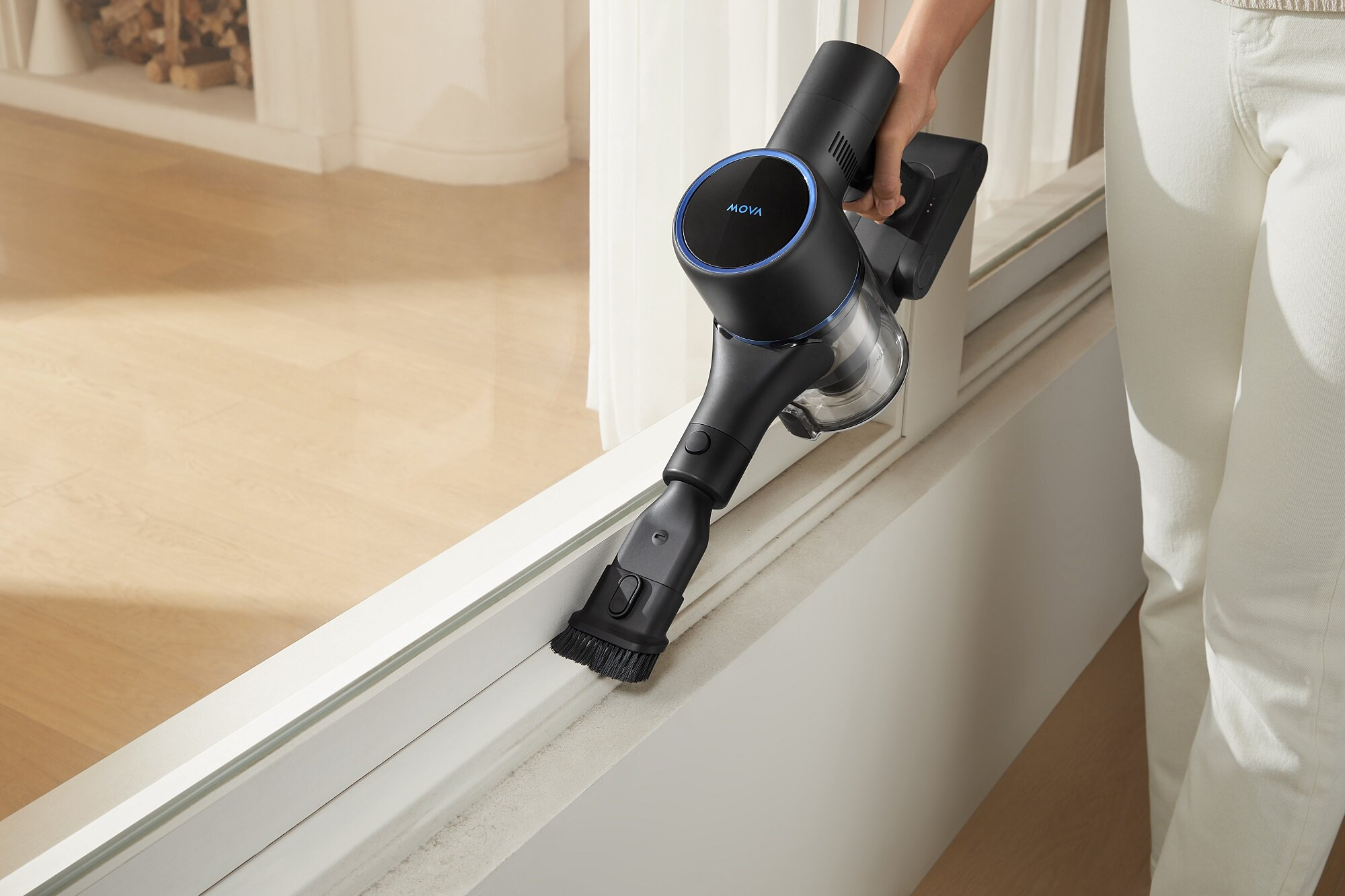 Osoba używa czarno-niebieskiego odkurzacza MOVA do czyszczenia ramy okiennej. Osoba ma na sobie białe spodnie.