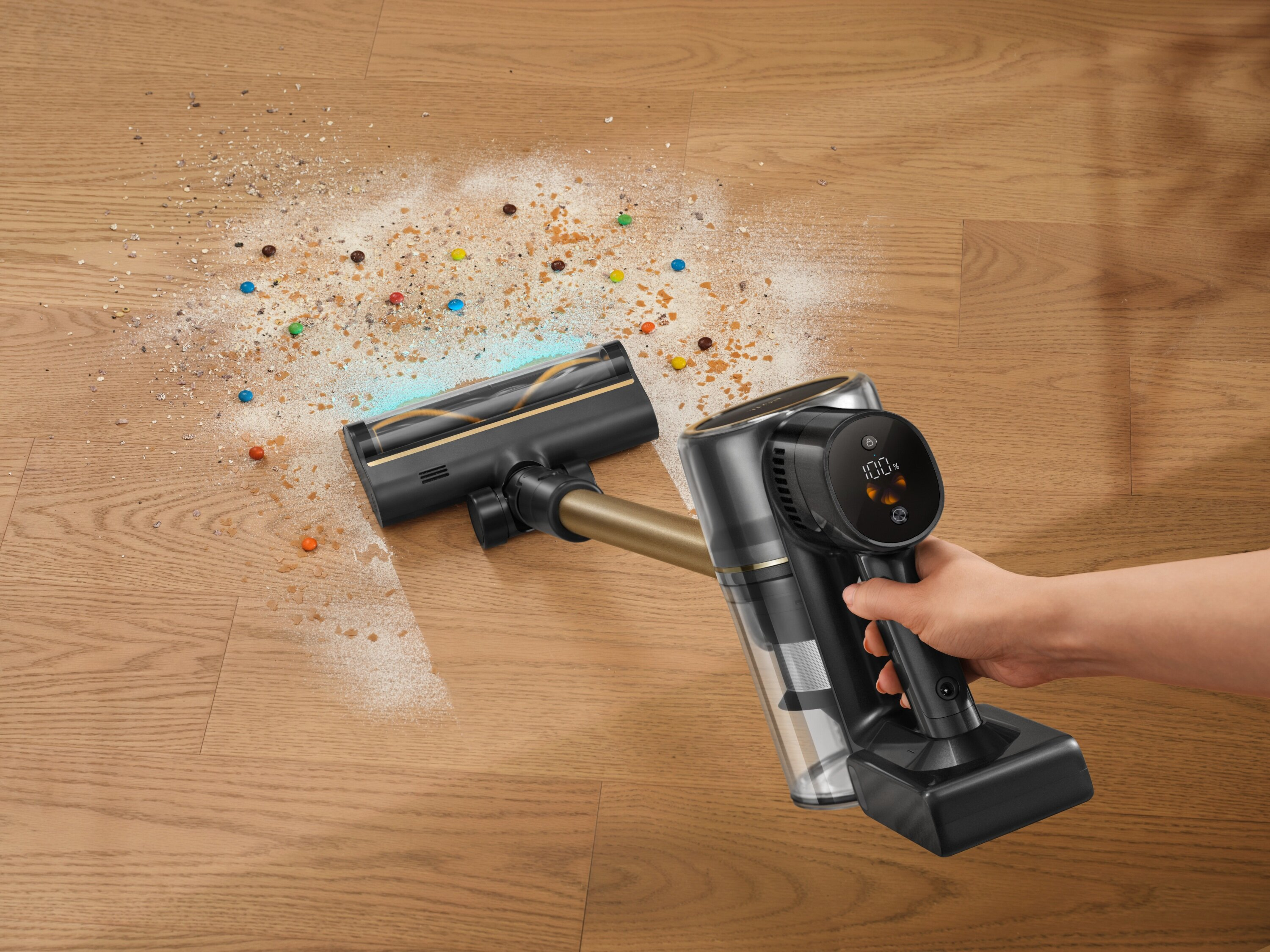 Odkurzacz bezprzewodowy sprząta rozsypane okruchy i cukierki na drewnianej podłodze. Wyświetlacz pokazuje 100%.