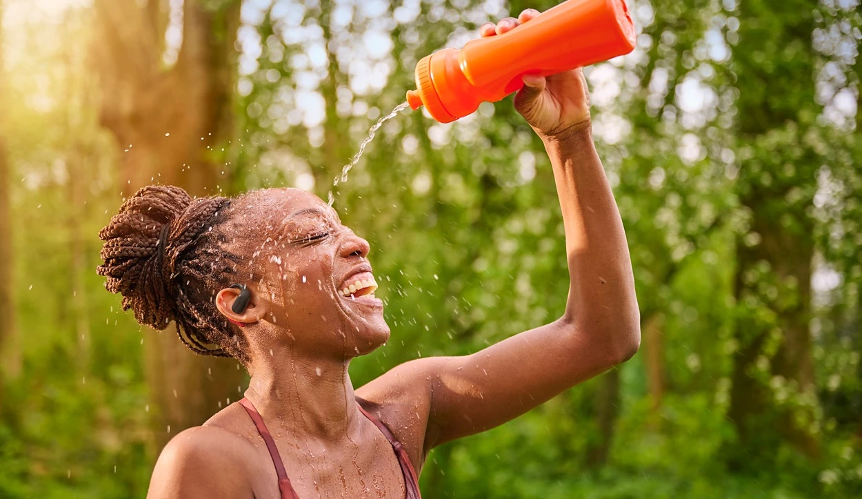 Kobieta polewa twarz wodą z pomarańczowej butelki, uśmiechając się. Ma na sobie biustonosz sportowy i upięte włosy. W tle zielone drzewa.