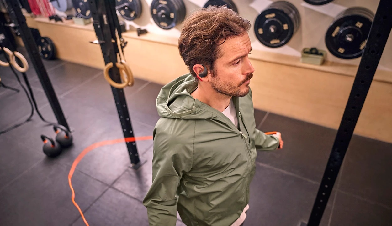 Mężczyzna na siłowni w słuchawkach i kurtce. Sprzęt, taki jak kółka, ciężary i kettlebells, jest w tle.