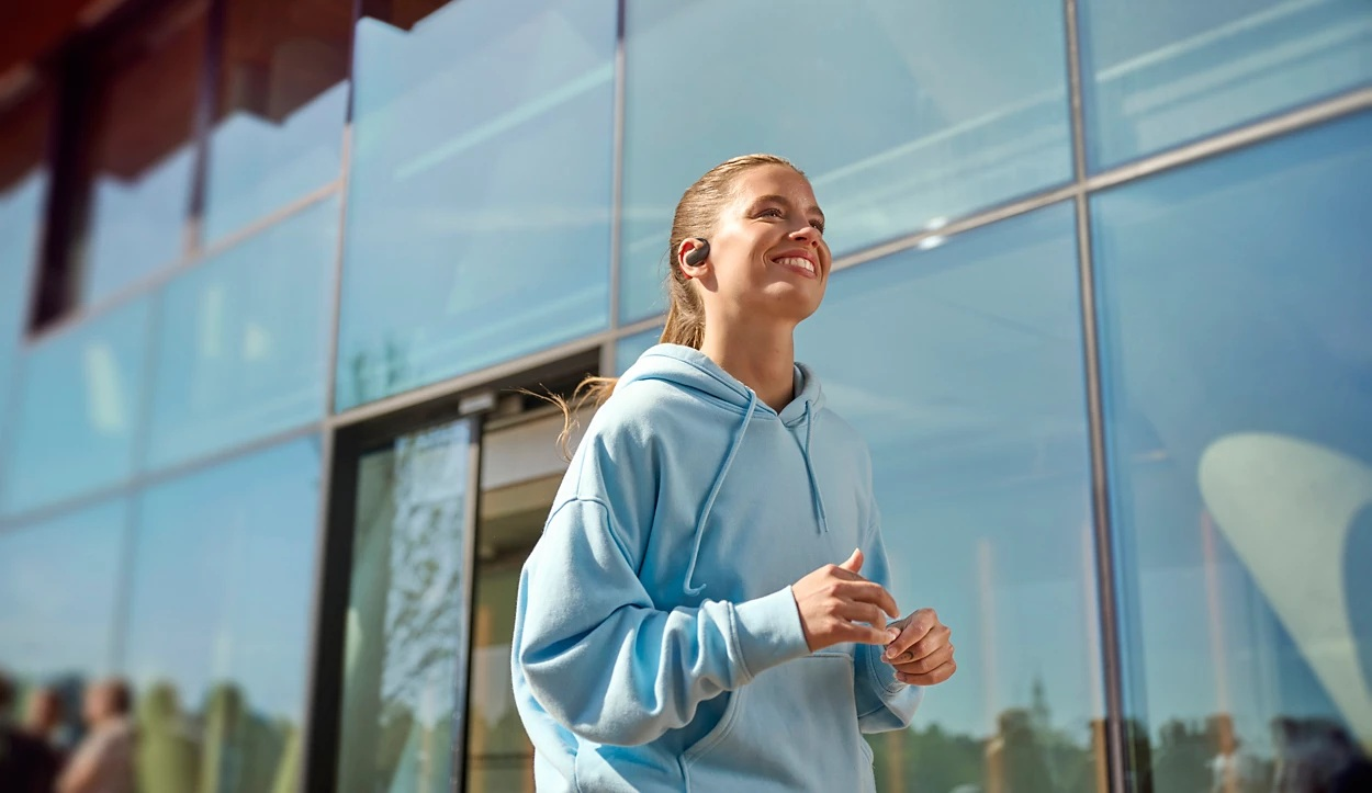 Uśmiechnięta kobieta w niebieskiej bluzie z kapturem i słuchawkami jest na zewnątrz. Tło obejmuje nowoczesny budynek ze szklanymi oknami.