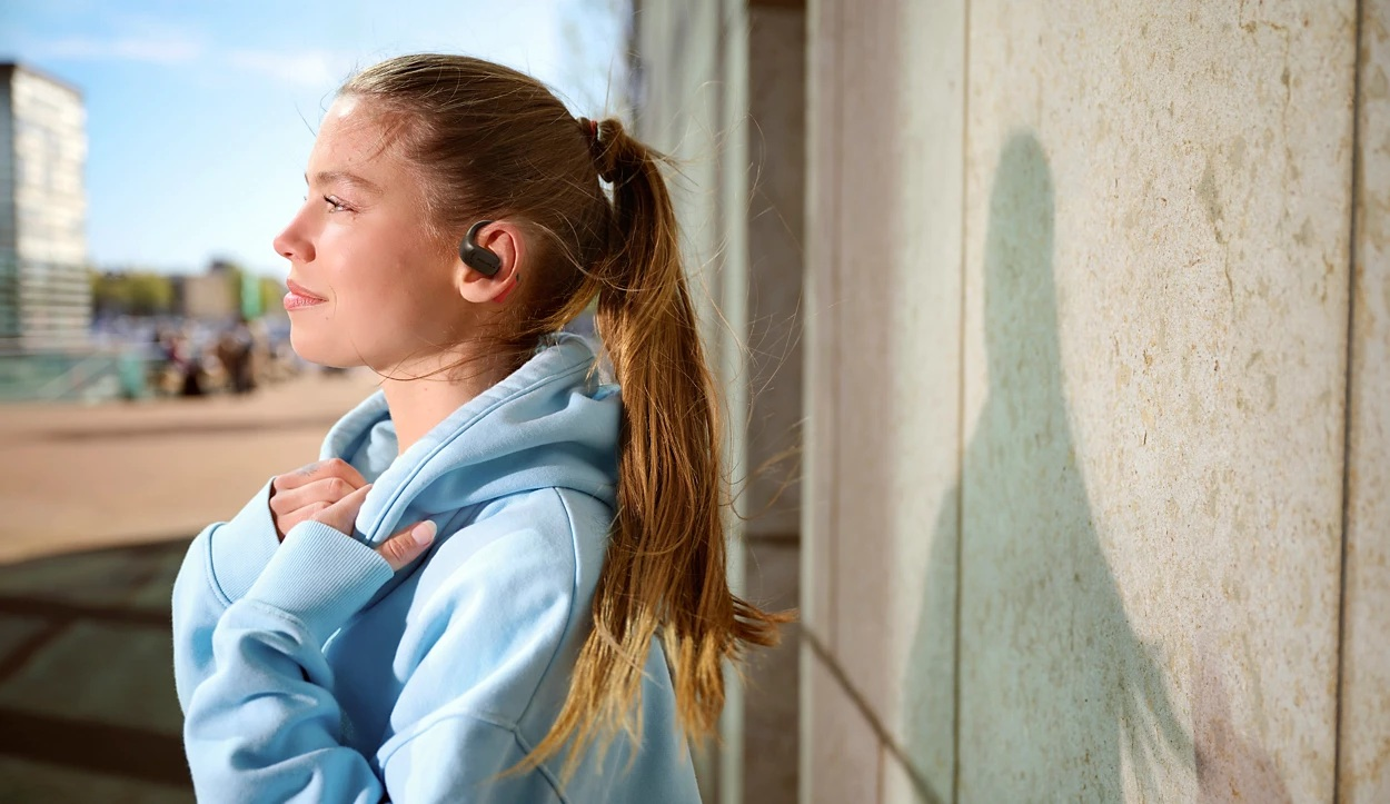 Młoda kobieta ze słuchawkami i niebieską bluzą z kapturem stoi na zewnątrz. Ma blond włosy spięte w kucyk, opiera się o kamienny mur.