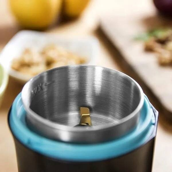 Zbliżenie na młynek. Kubek ze stali nierdzewnej ze złotym ostrzem. Niebieski pierścień wokół kubka. Rozmyte produkty spożywcze w tle.