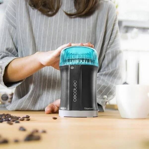 Osoba trzyma młynek do kawy z niebieską pokrywką. Ziarna kawy i kubek znajdują się w pobliżu na drewnianej powierzchni.