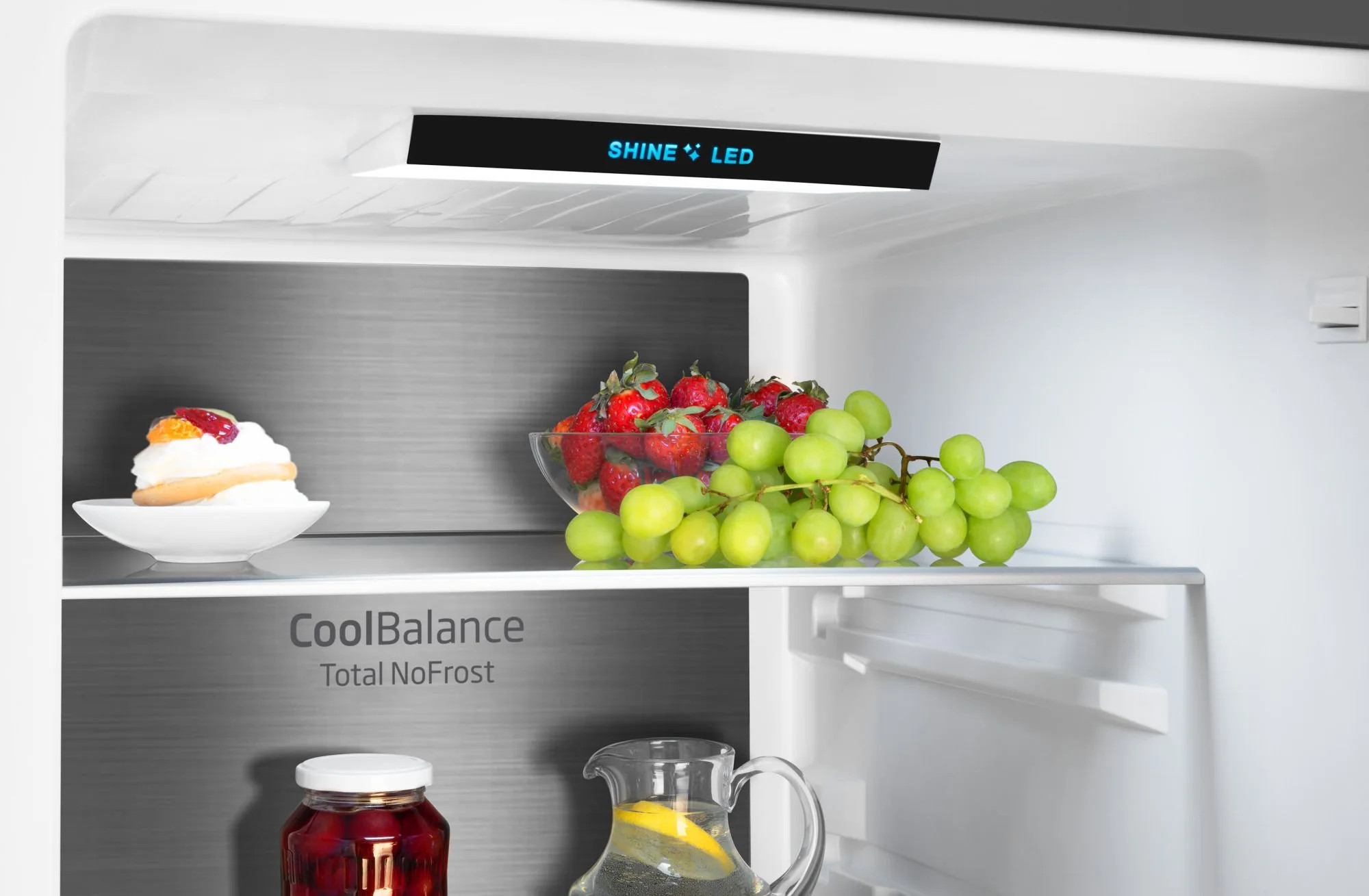 Wewnątrz lodówki: owoce, ciasto i słoik. Truskawki i winogrona na półce. Światło LED Shine. Tekst CoolBalance na tylnej ścianie.