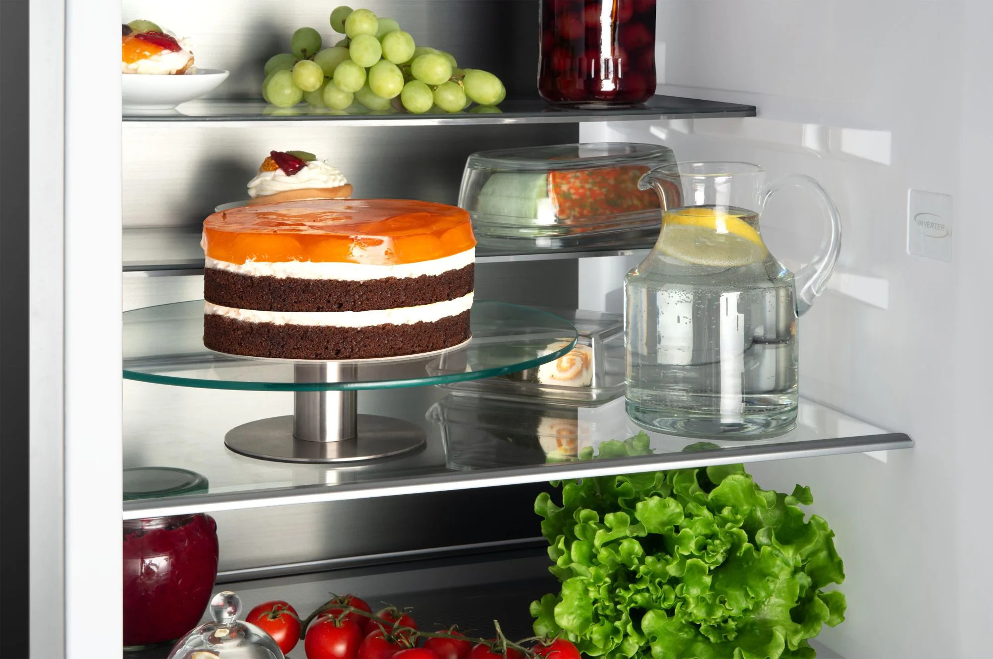 Wewnątrz lodówki widać ciasto, winogrona i dzbanek z wodą. Inne przedmioty to sałata, pomidory i słoik wiśni.