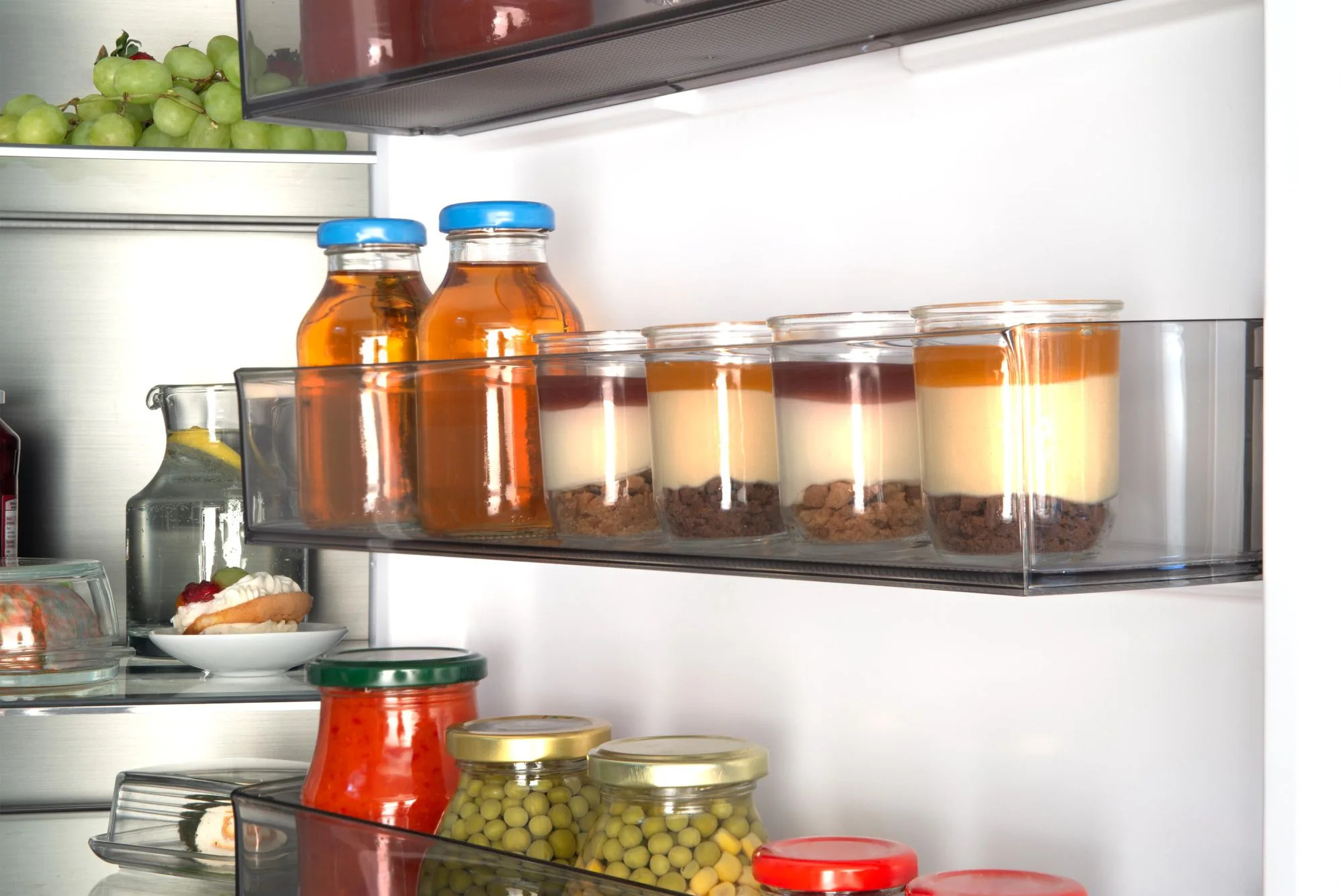 Wnętrze lodówki z butelkami soku, deserami, słoikami z groszkiem i sosem oraz winogronami. Wyświetlane są różne produkty spożywcze.