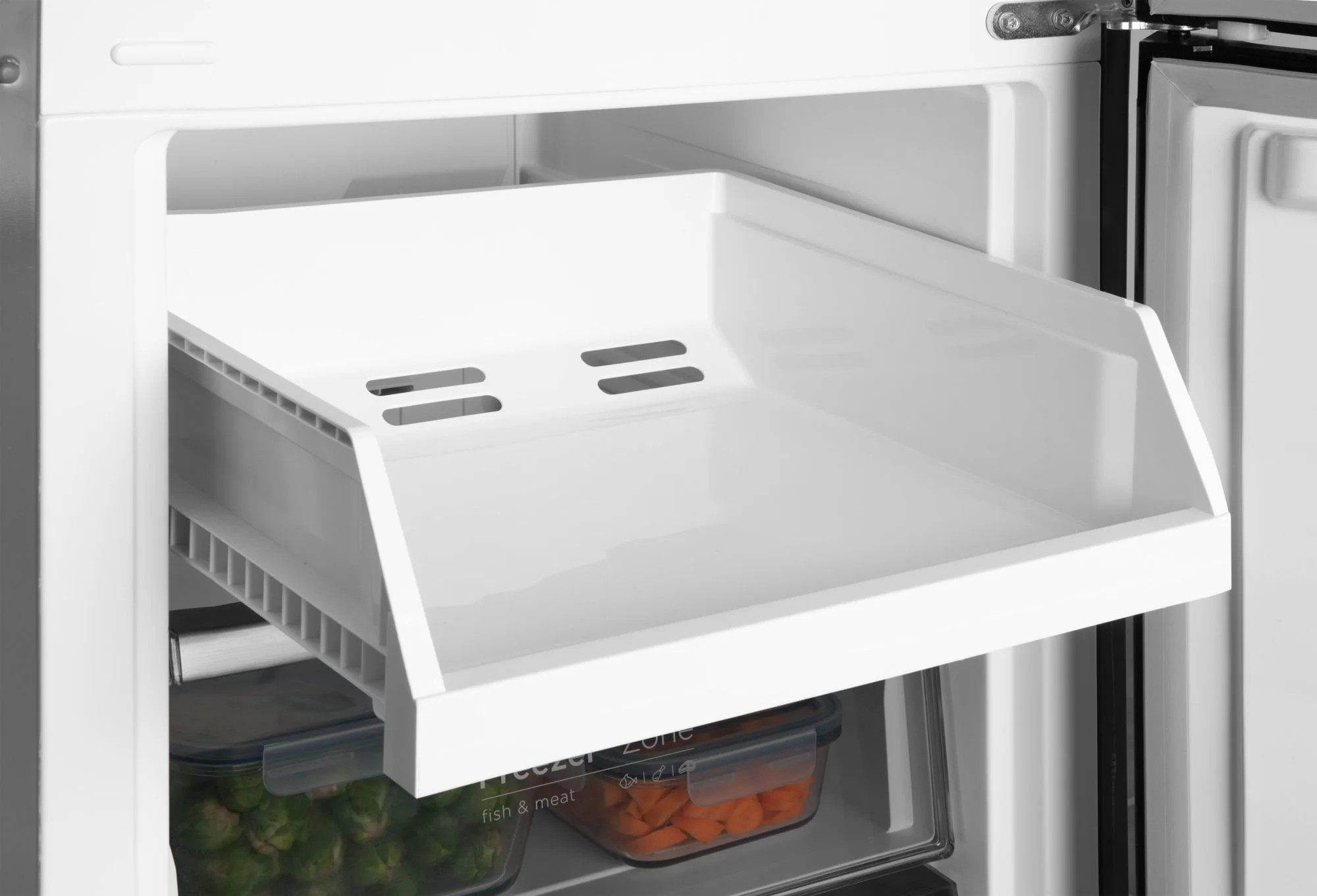 Otwarta lodówko-zamrażarka. Biała plastikowa półka jest wysunięta. Poniżej pojemniki z warzywami i marchewką. Strefa zamrażania.