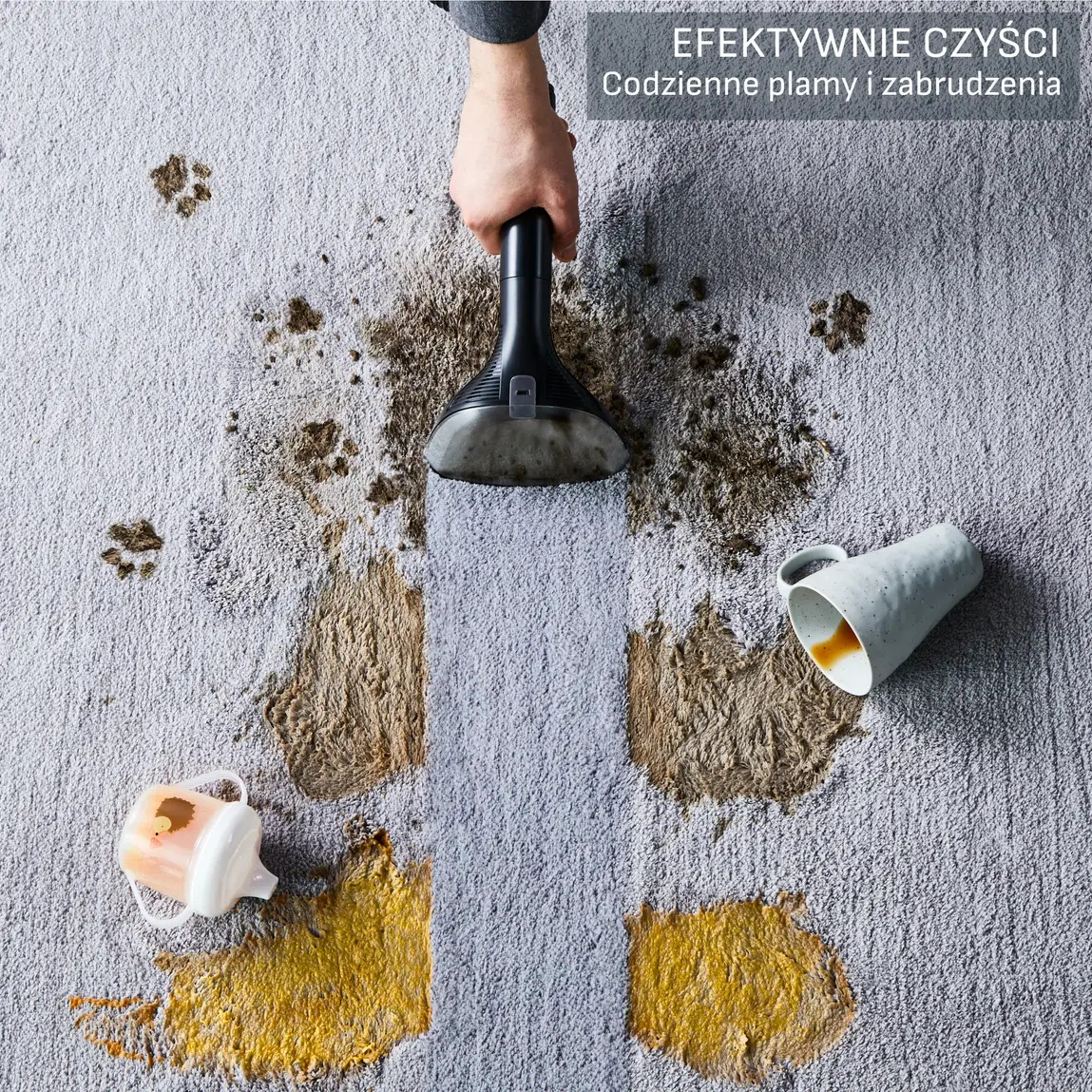 Dywan jest poplamiony rozlanymi płynami i śladami łap. Ręka trzyma odkurzacz nad oczyszczonym paskiem. Kubki są przewrócone.
