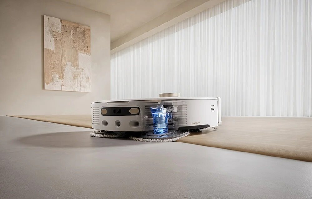 Biały odkurzacz robot stoi na jasnej drewnianej powierzchni. Obraz wisi na ścianie. Tło: biała zasłona.