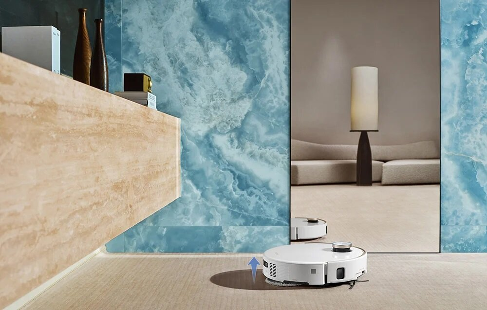 Robot sprzątający na dywanie, odbijający się w lustrze. Tło przedstawia sofę, lampę i niebieską marmurową ścianę.