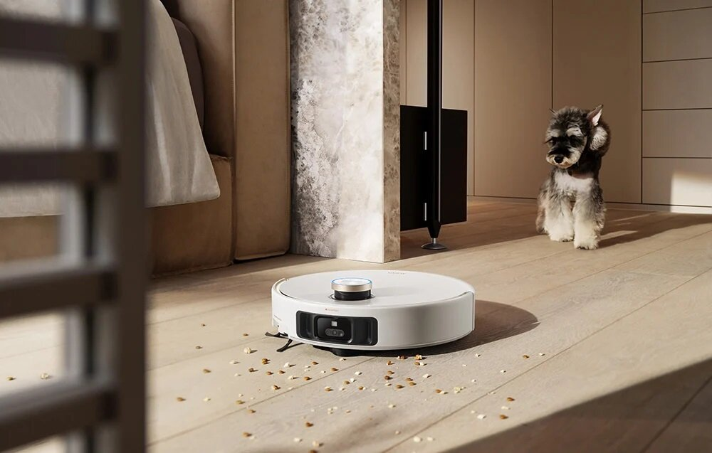 Biały robot sprząta drewnianą podłogę. W pobliżu siedzi mały pies. Na podłodze leżą śmieci. W tle znajduje się łóżko.