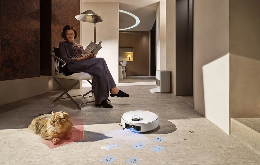 Kobieta czyta, kot leży, robot sprzątający na podłodze. Nowoczesne wnętrze z jasnymi i ciemnymi tonami.