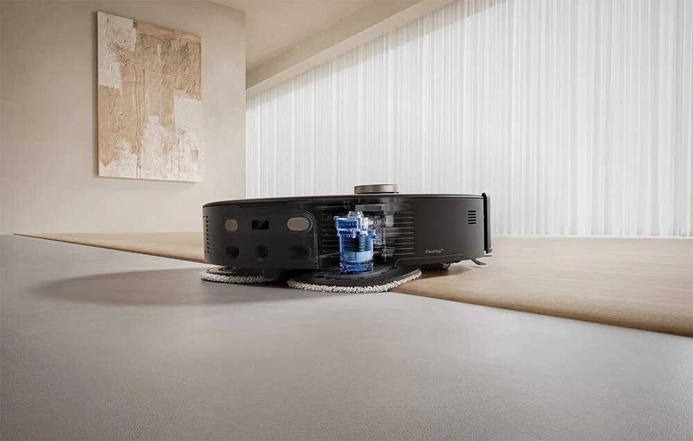 Czarny robot odkurzający na drewnianej podłodze. Obraz wisi na ścianie. W tle zasłony.