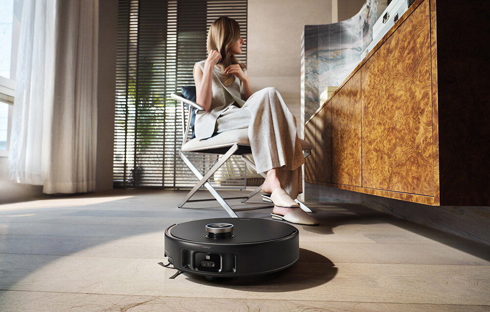 Kobieta siedzi, a robot sprząta. Po prawej stronie drewniana szafka. Światło słoneczne prześwituje przez żaluzje.