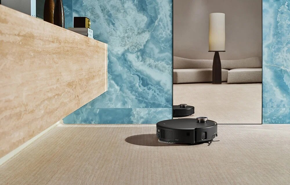 Czarny robot odkurzający na beżowym dywanie. Lustro odbija lampę i sofę. Niebieska marmurowa ściana w tle.
