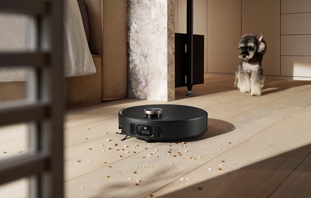 Czarny odkurzacz automatyczny na drewnianej podłodze z zanieczyszczeniami. W tle stoi pies. Widoczne jest również łóżko i dekoracyjna ściana.