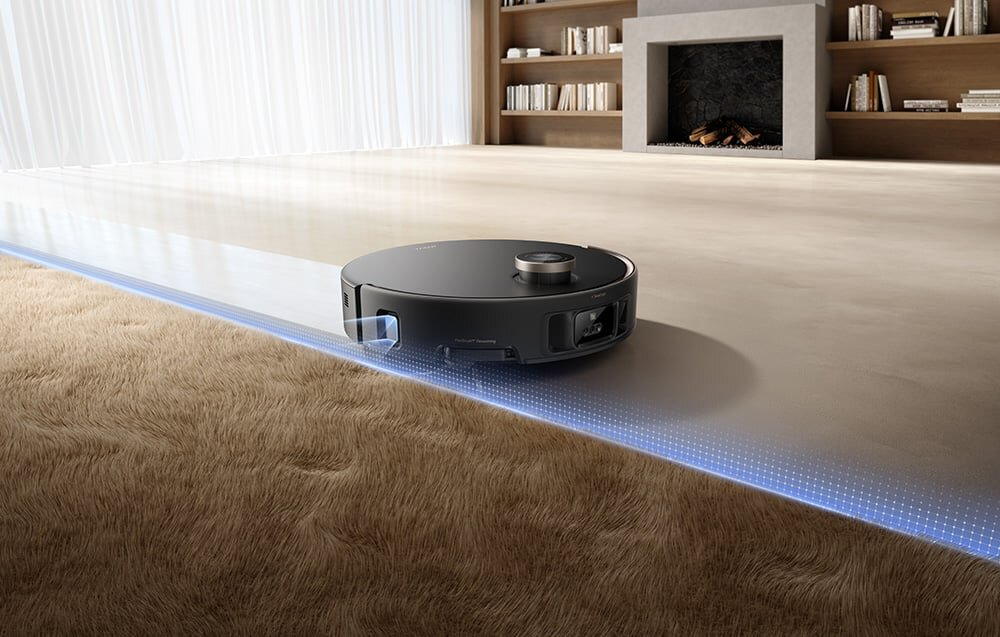 Czarny robot sprzątający na dywanie. Niebieska przerywana linia wskazuje ścieżkę czyszczenia. Tło przedstawia salon z kominkiem.