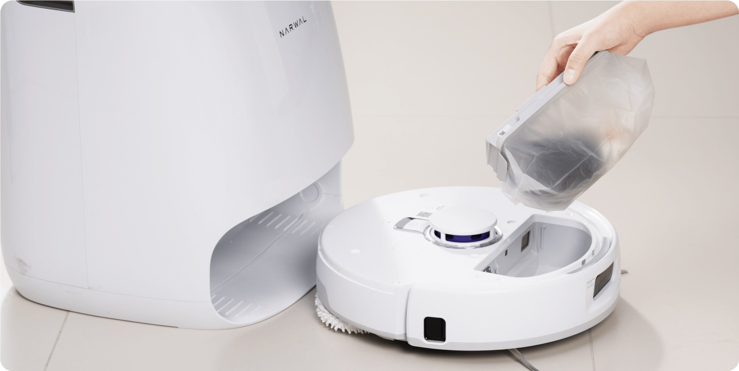 Biały odkurzacz robot Narwal. Ręka trzyma worek, wlewając zawartość do otworu odkurzacza. Biała podstawa znajduje się po lewej stronie.