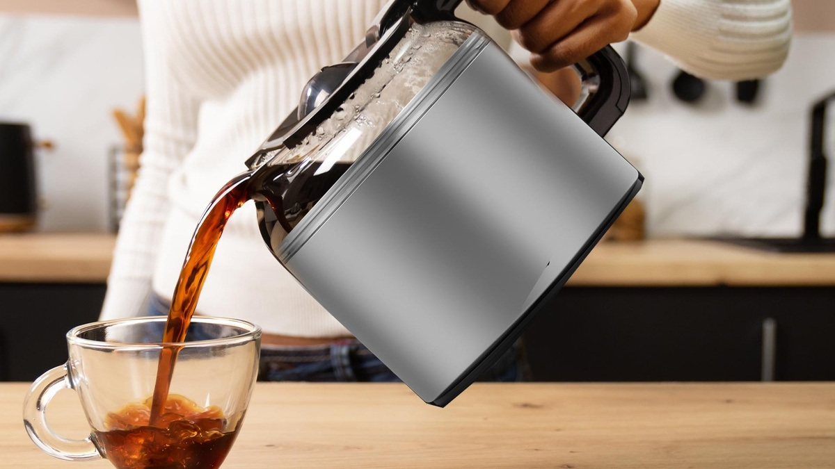 Osoba nalewa kawę ze srebrnej karafki do przezroczystego szklanego kubka. Widoczny strumień kawy. Tło przedstawia kuchnię.