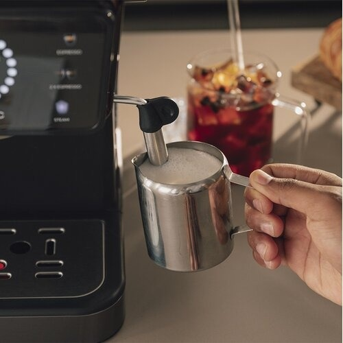Osoba spienia mleko ekspresem do kawy. Napój z owocami w tle. Ręka trzyma metalowy dzbanek.