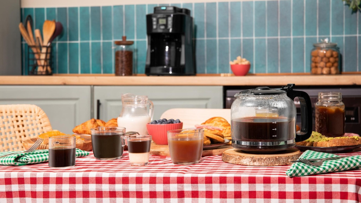 Stół śniadaniowy z kawą, ciastkami i owocami. W tle ekspres do kawy. Stół ma czerwoną i białą kratowaną obrus.