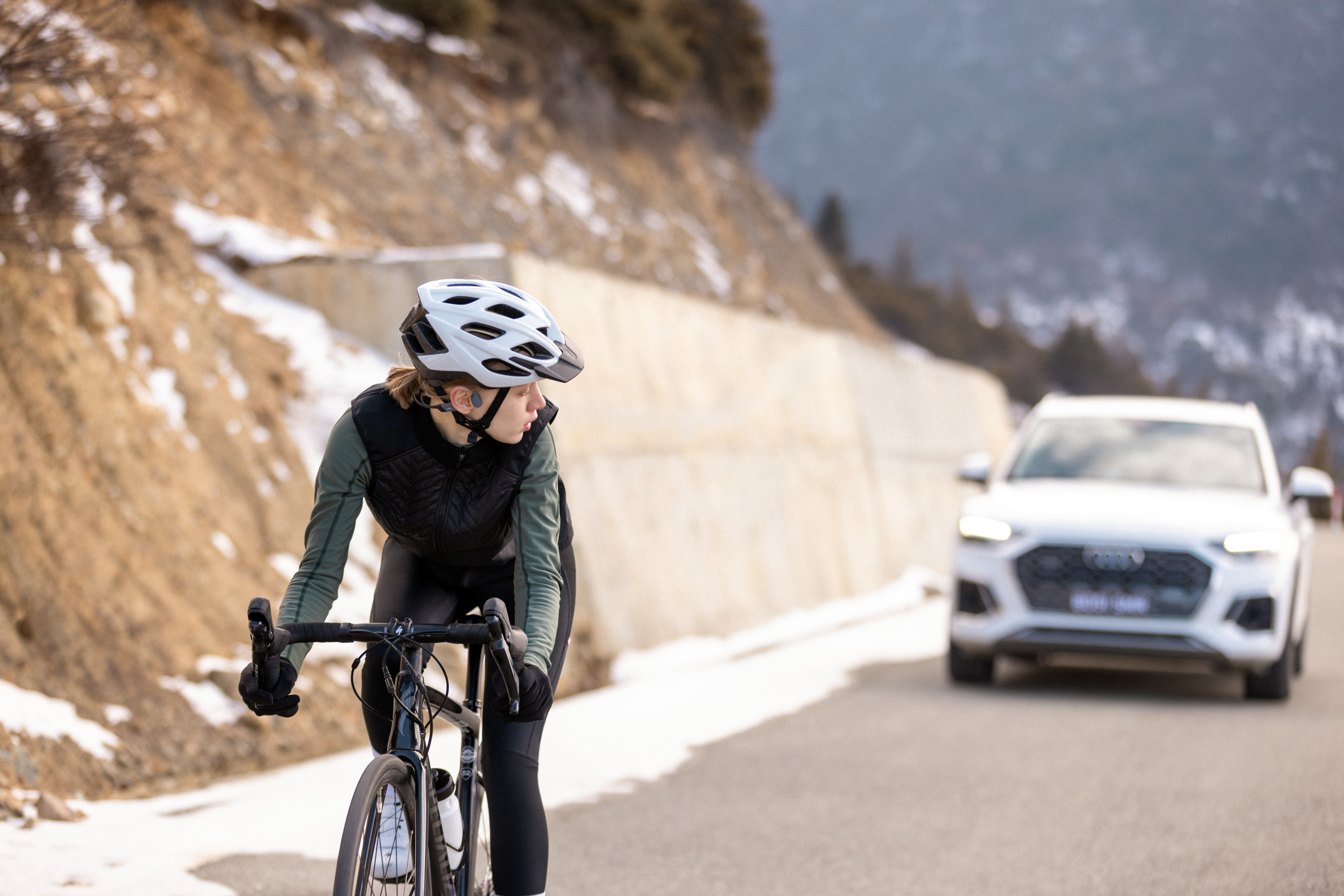 Radfahrer auf der Straße mit Helm. Ein weißer SUV im Hintergrund. Verschneiter Berg und brauner Hügel sichtbar.