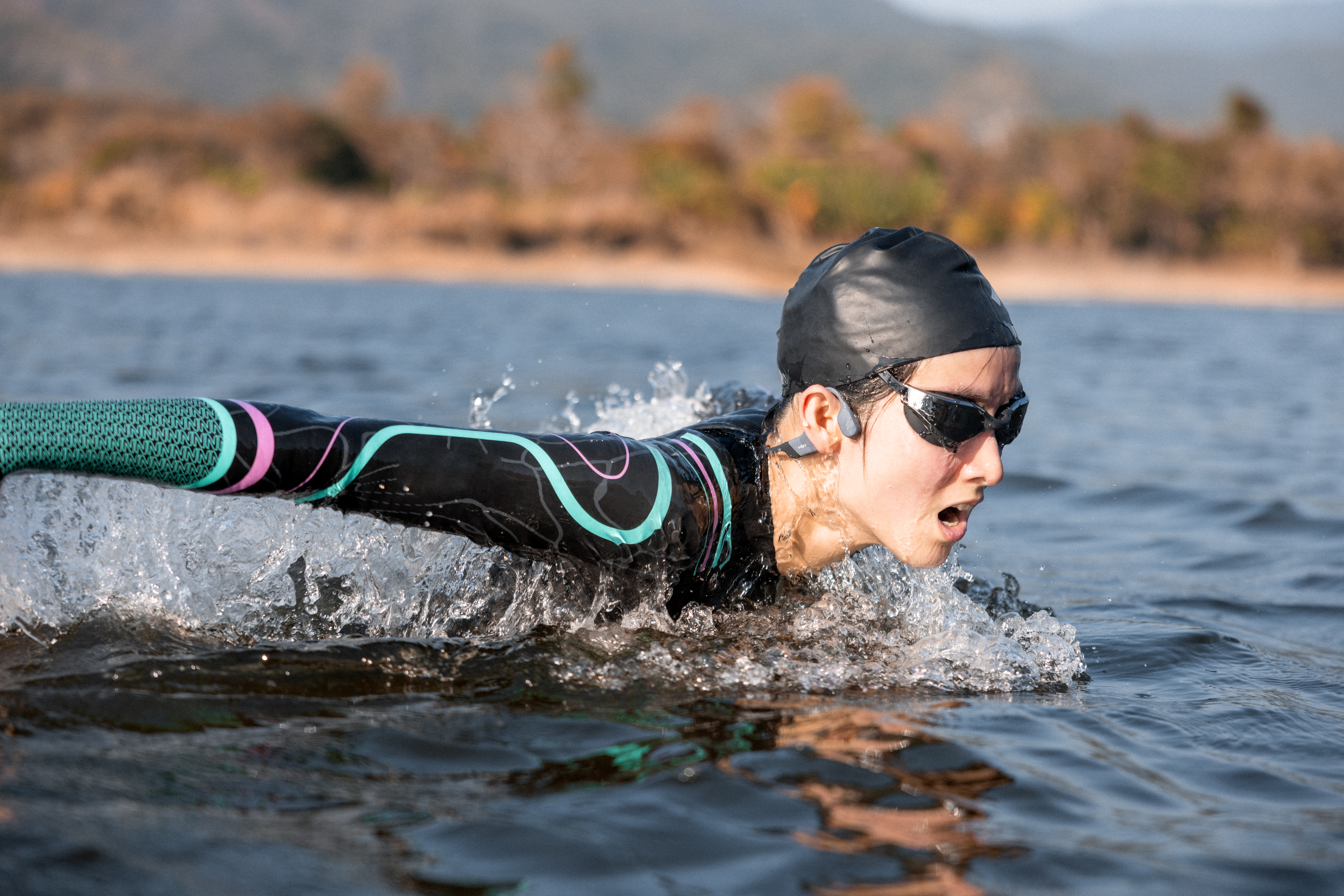 Eine Schwimmerin mit schwarzer Kappe und Brille schwimmt in einem See. Wasser spritzt um sie herum. Hintergrund zeigt Wasser und Bäume.