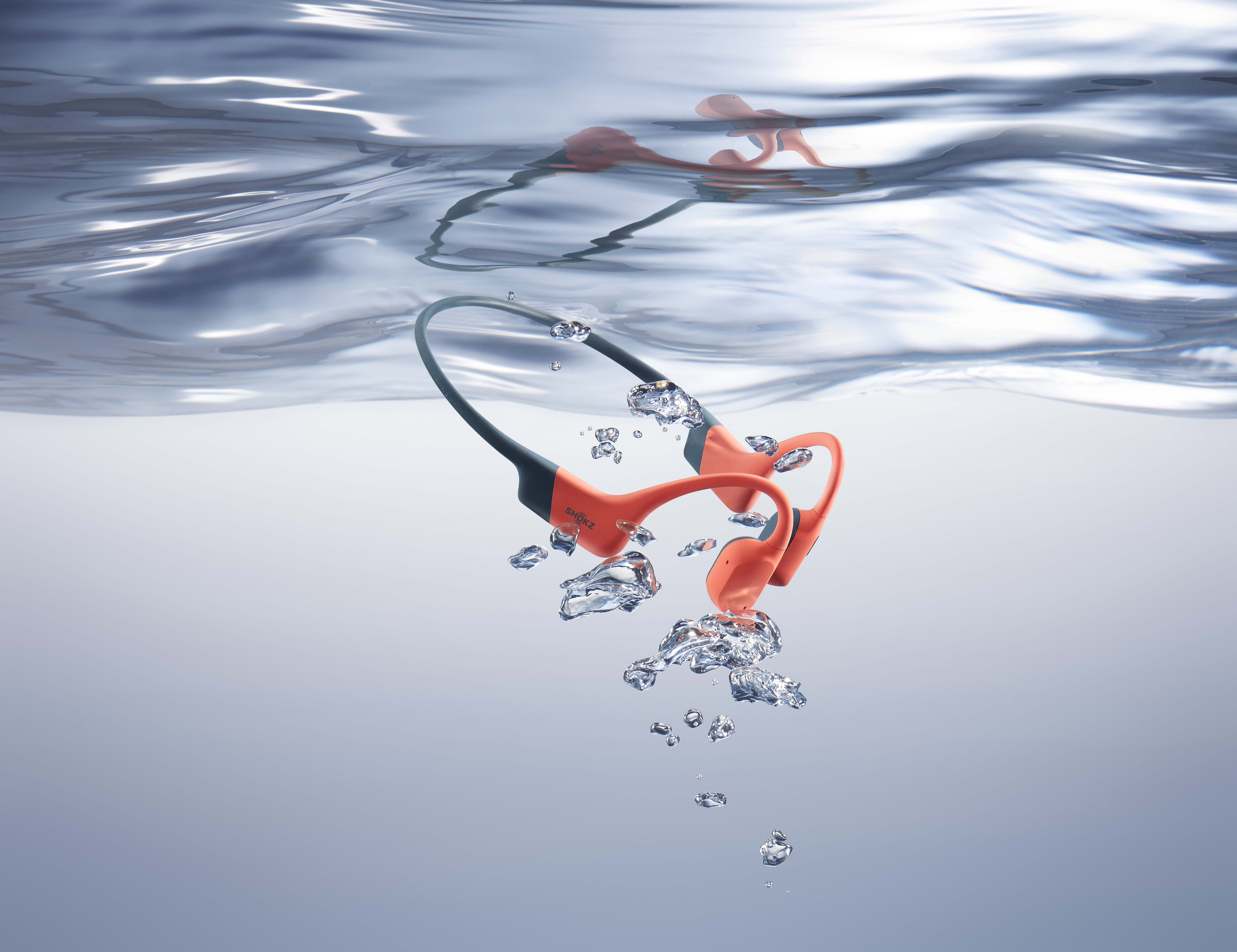 Orange-graue Knochenschallkopfhörer unter Wasser. Blasen umgeben die Kopfhörer. Wasseroberfläche ist oben sichtbar.