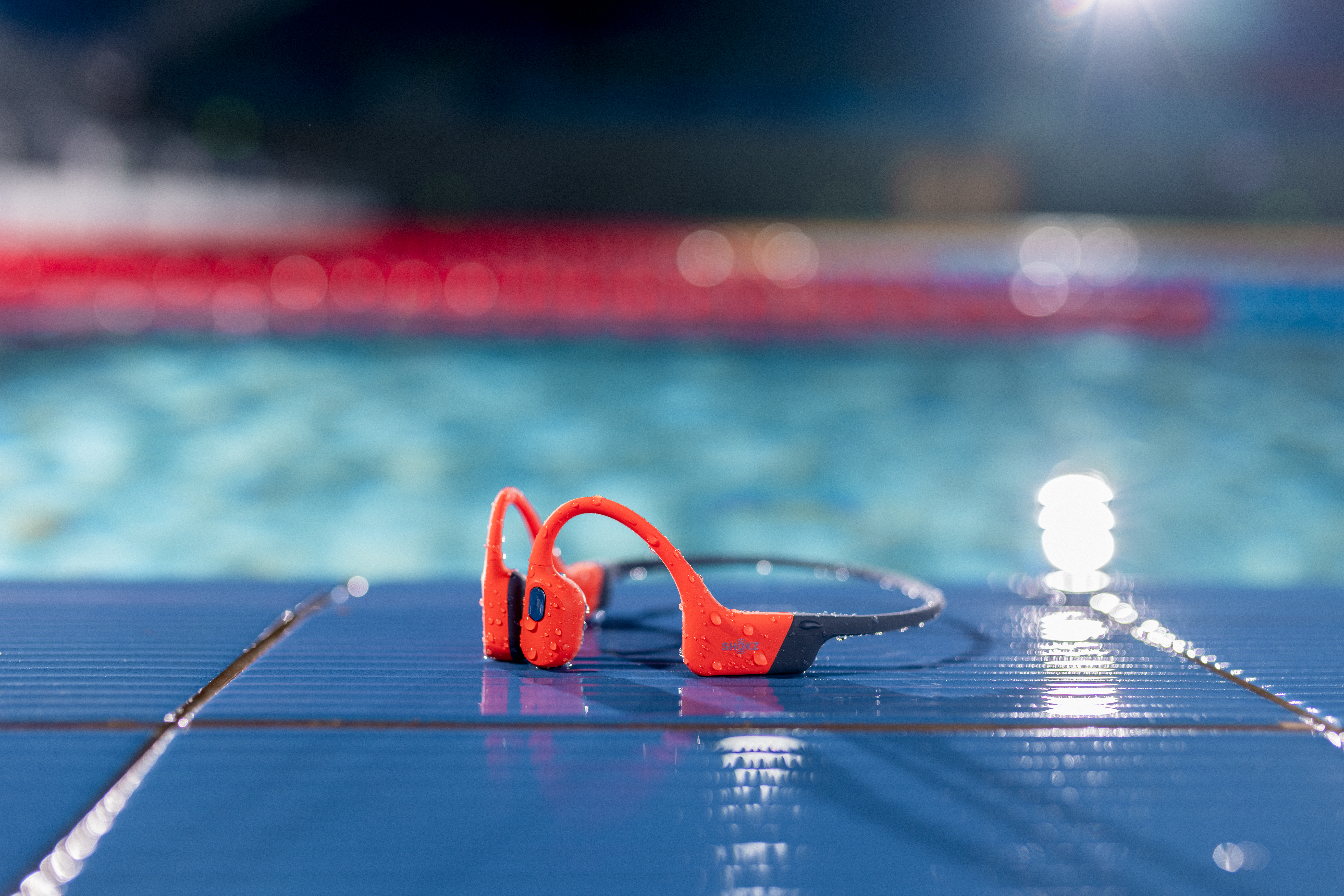 Orange-schwarze Kopfhörer mit Wassertropfen liegen auf einer blauen Oberfläche. Hintergrund ist ein Pool mit roten Bahnen.