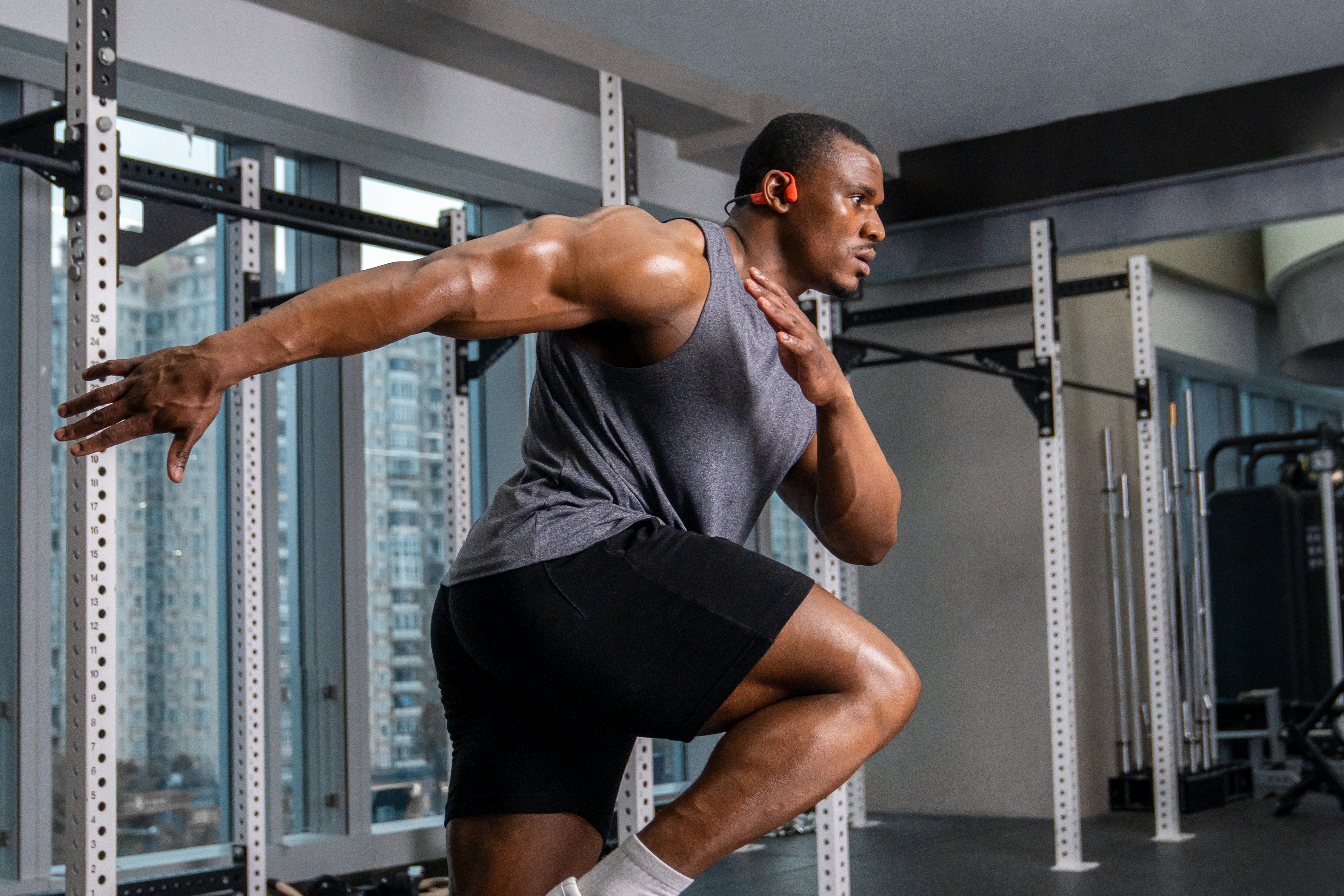 Muskulöser Mann im Fitnessstudio läuft, trägt ein graues Oberteil und schwarze Shorts, mit orangefarbenen Kopfhörern. Fitnessgeräte und Stadtblick im Hintergrund.