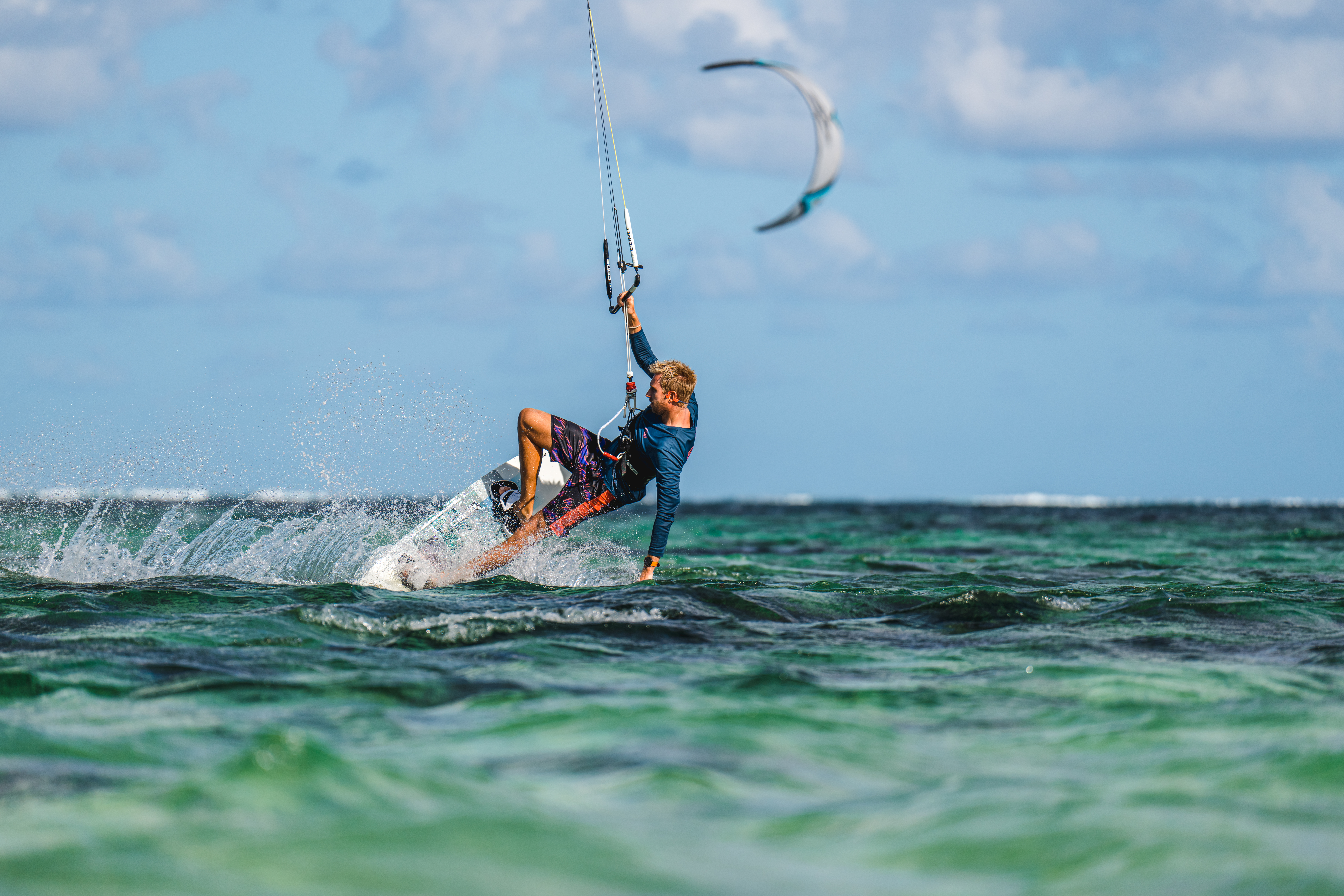 Kitesurfer auf einem Board in türkisfarbenem Wasser, mit einem Kite am Himmel. Die Person trägt ein blaues Hemd und bunte Shorts.