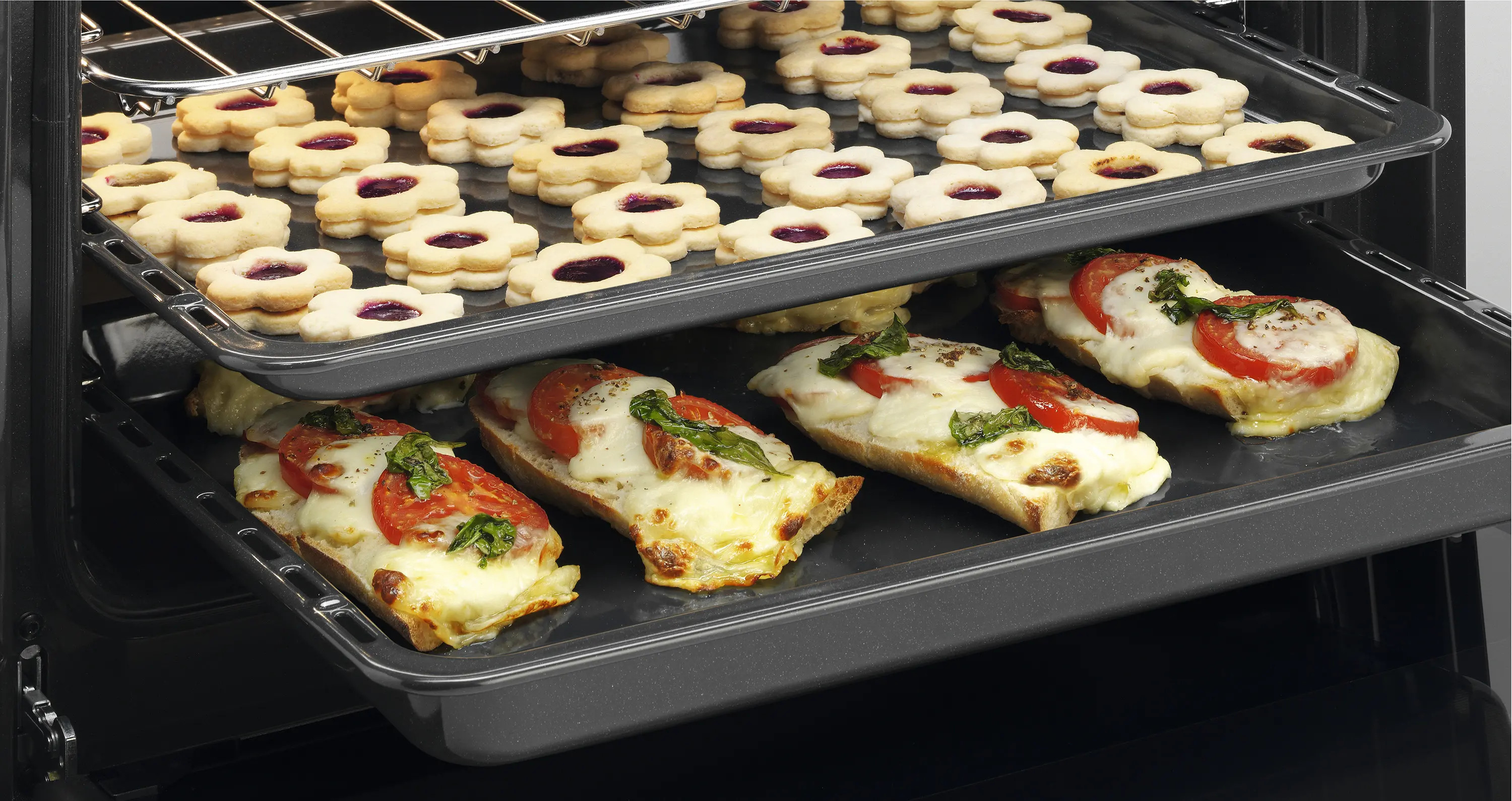 Piekarnik z dwiema tacami. Górna taca z ciasteczkami w kształcie kwiatów z dżemem. Dolna taca z chlebem z pomidorami, mozzarellą i bazylią.