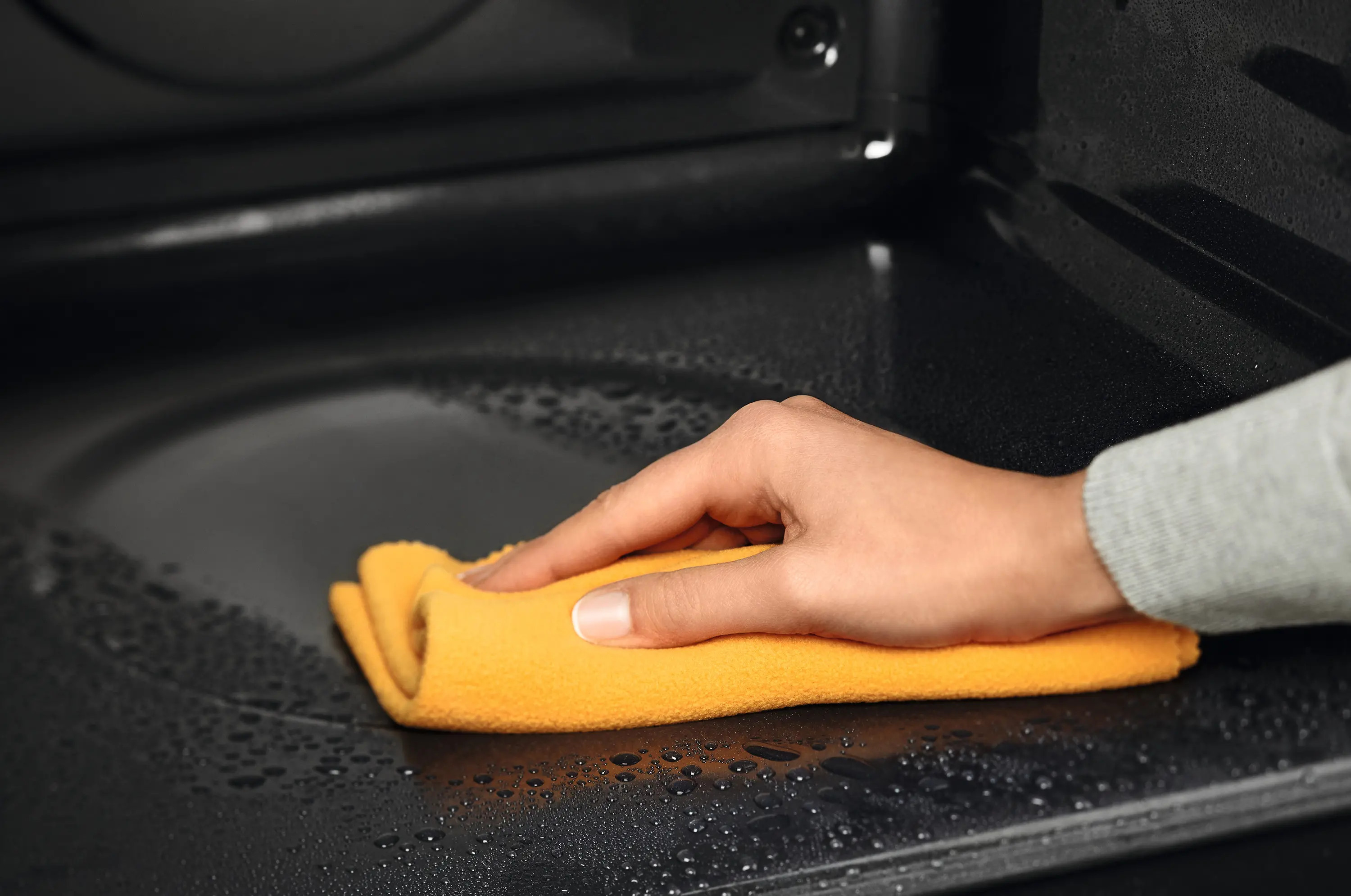 Ręka z żółtą szmatką czyści czarną powierzchnię. Powierzchnia ma krople wody, prawdopodobnie w kuchence mikrofalowej.