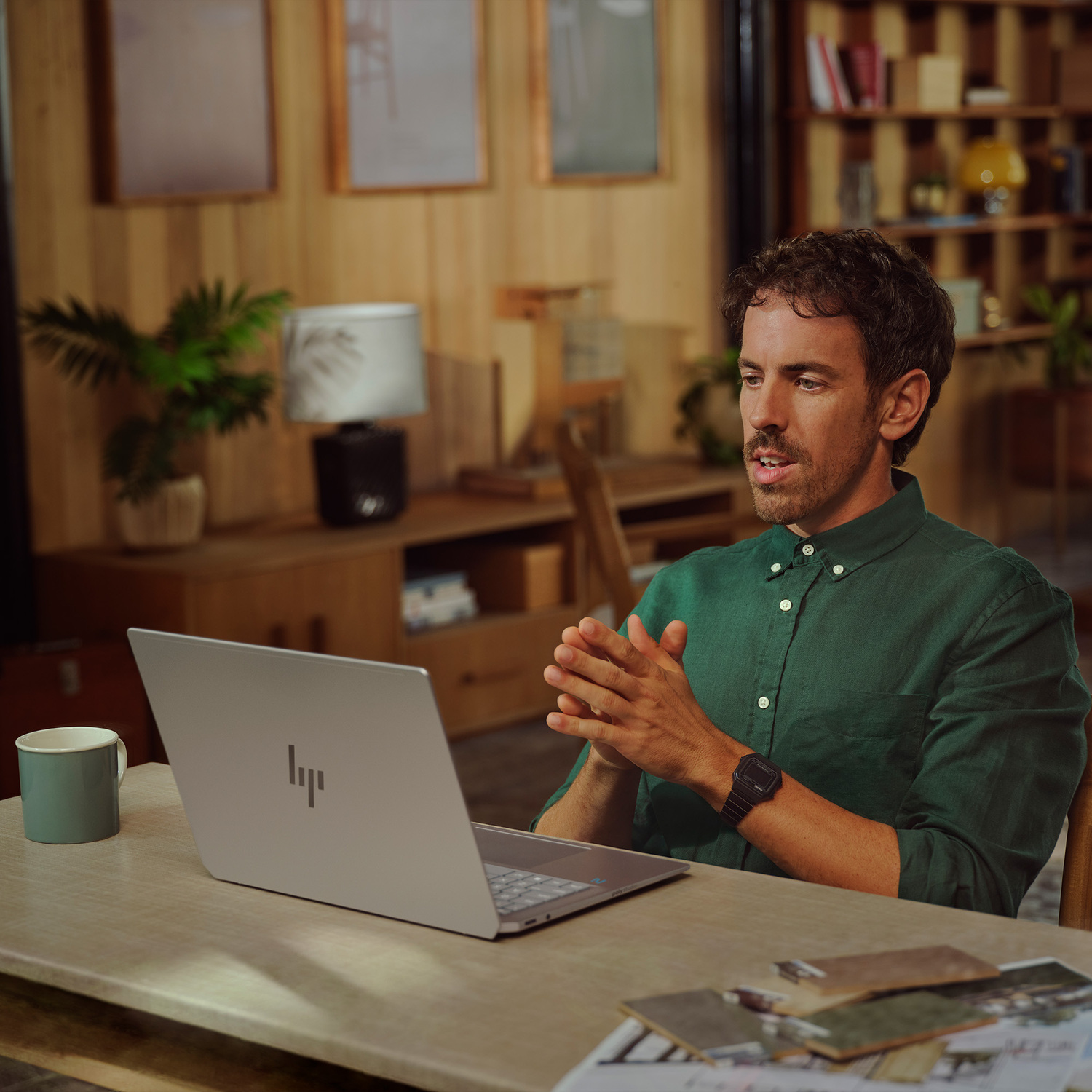 Man in green shirt using a laptop. Hands clasped. Mug and laptop on table. Wooden backdrop and shelves in the background.