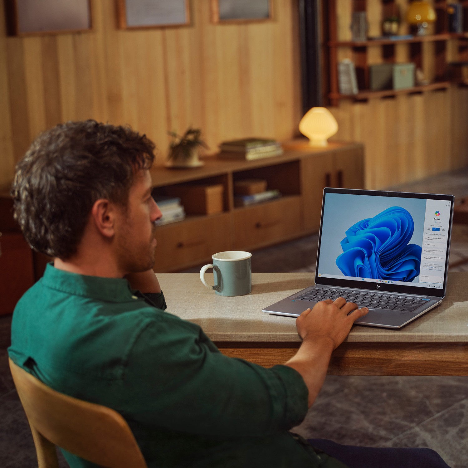 A man in a green shirt is using a laptop at a wooden table. A mug sits to the left. The screen shows a blue design.